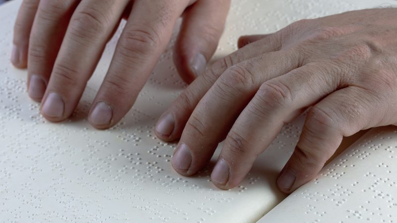two hands rest on a line of Braille in a bound Braille book.